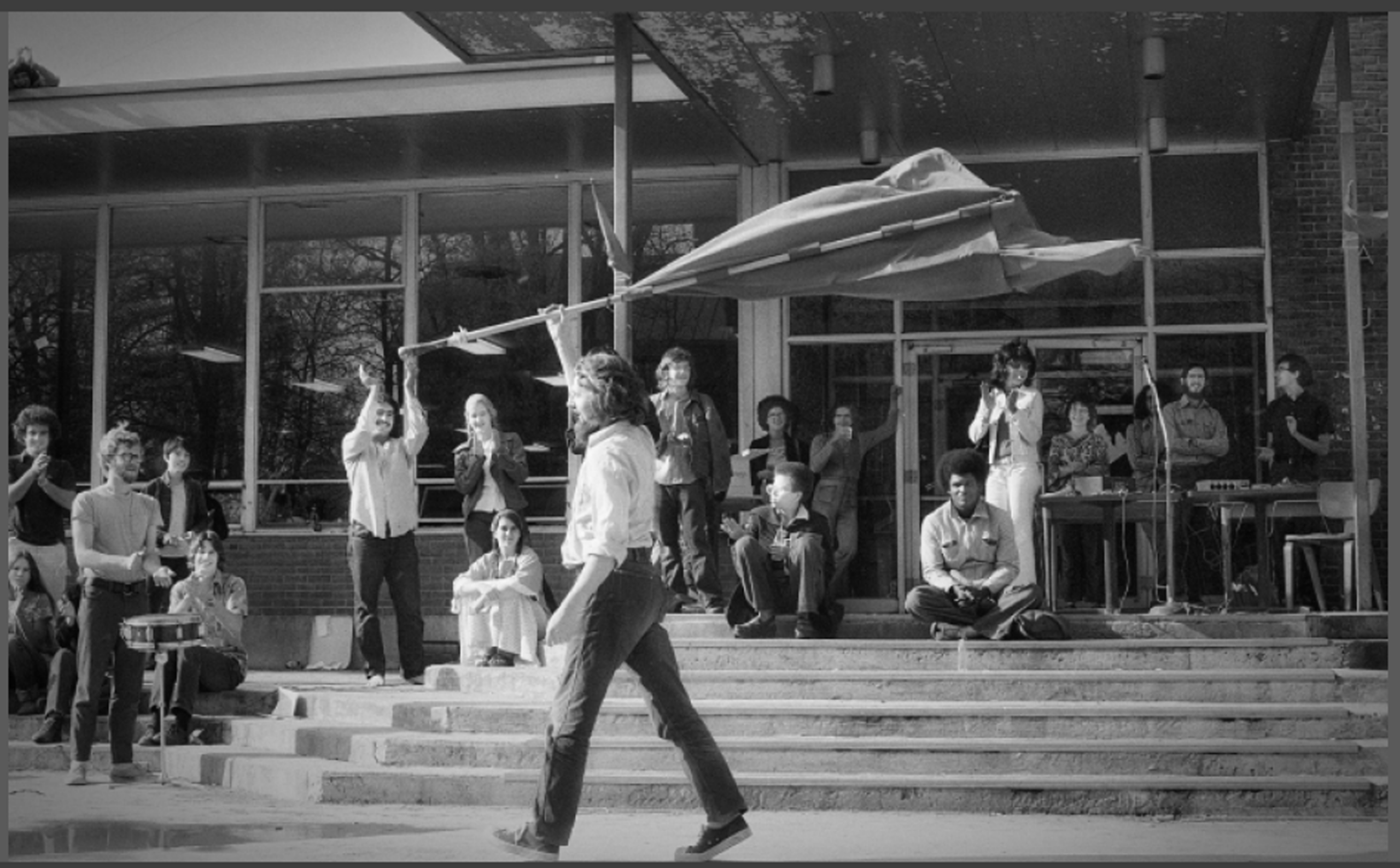 Students sitting on Union Building stoop steps applauding another student who is marching by waving a flag.